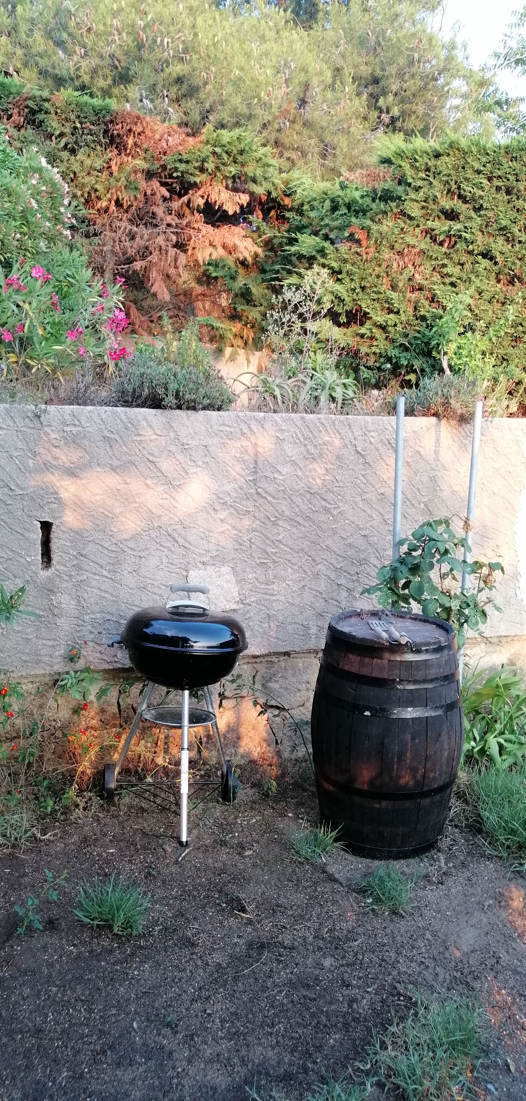 Een zwarte barbecue met deksel en een metalen standaard, een houten vat, en een stenen muur met begroeiing op de achtergrond.