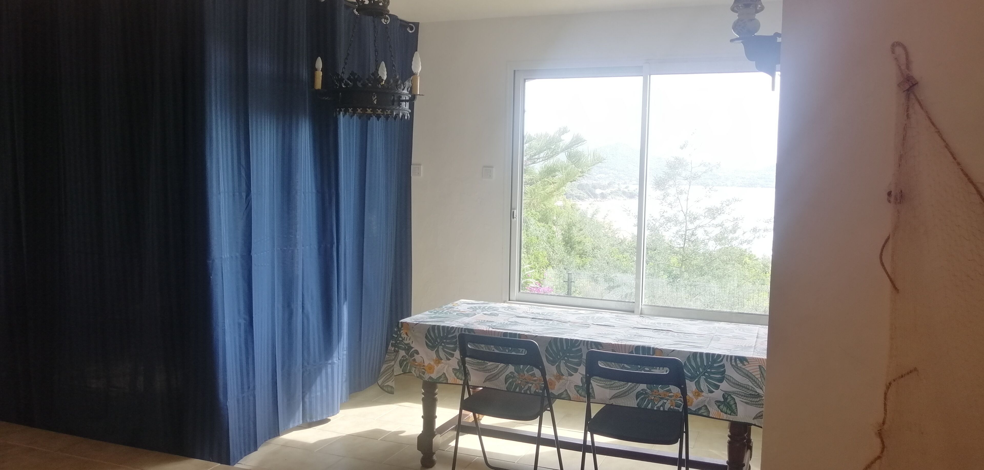 A dining area with a table and chairs, a large window with a view of greenery, and blue curtains.