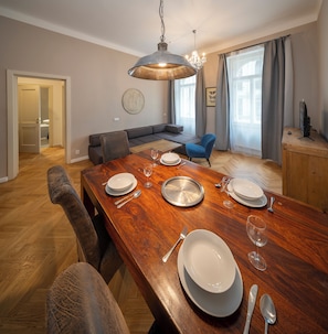 Un comedor con una mesa de madera dispuesta para cuatro personas, una araña y un espacio de sala moderno con un sofá y una televisión.