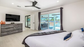 Ein modernes Schlafzimmer mit Bett, Fernseher, Deckenventilator und einer Glasschiebetür, die auf eine Terrasse mit Gartenmöbeln führt.