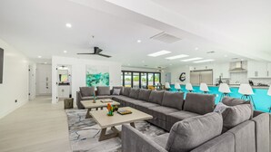Ein modernes Wohnzimmer mit einem großen, grauen Ecksofa, einem Holztisch und Blick auf die Küche sowie den Poolbereich.