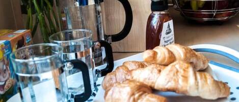 A kitchen counter with a coffee maker, a bottle of syrup, a plate with croissants, and a fruit basket.