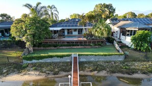 A dock extending into a calm body of water with a residential area in the background, including houses with solar panels and a well-maintained lawn. - River front property, huge yard and pool, 2km from the beach.  (Mermaid Waters)