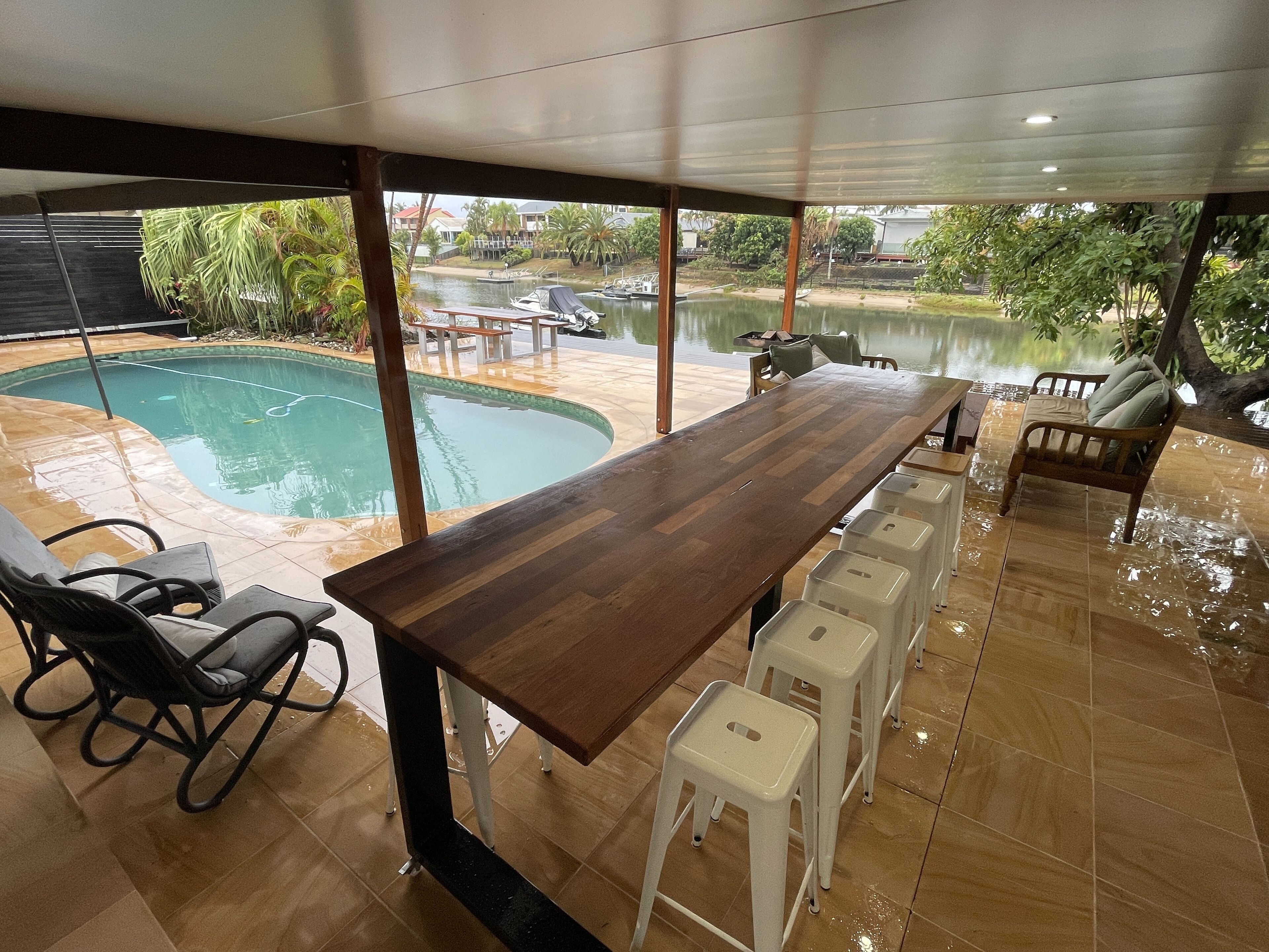 A dining area with a long wooden table, white stools, and a view of a swimming pool and boats.