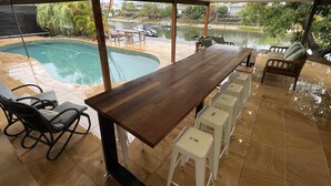 A dining area with a long wooden table, white stools, and a view of a swimming pool and boats. - River front property, huge yard and pool, 2km from the beach.  (Mermaid Waters)