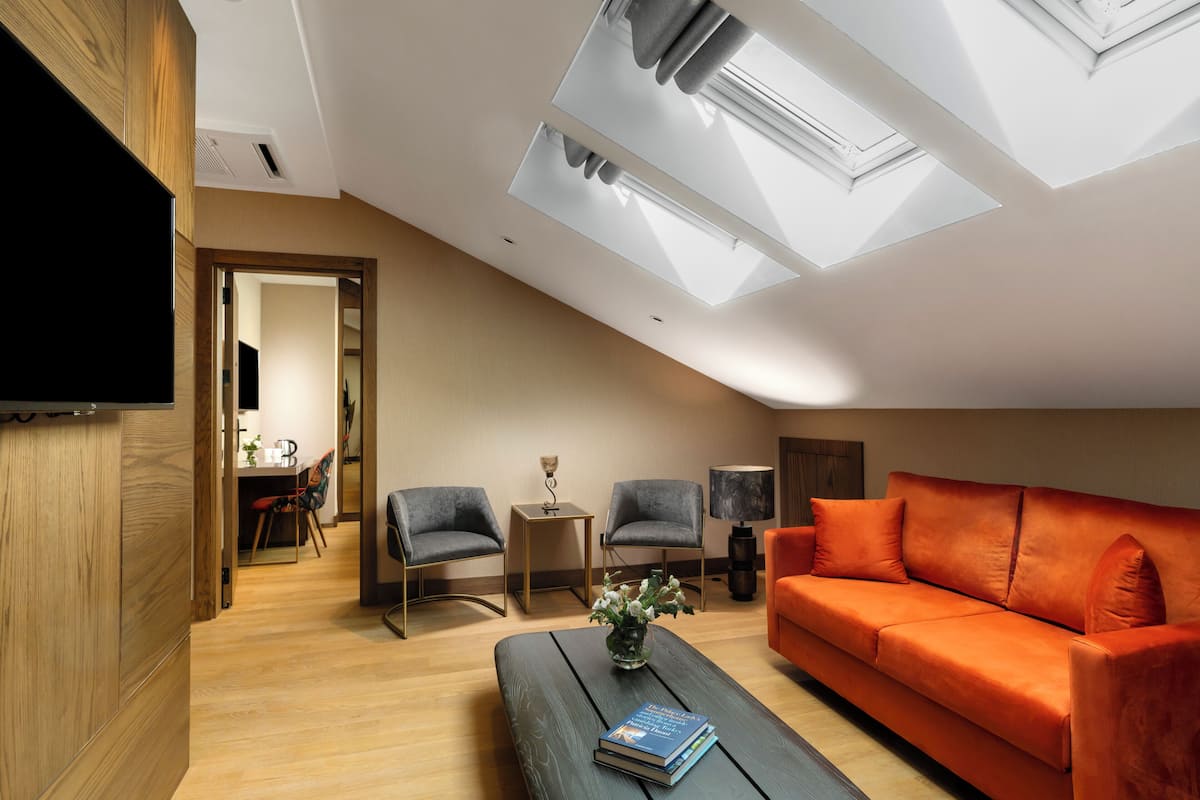 A modern living room with a sloped ceiling, skylight, orange sofa, two armchairs, a coffee table with books, and a side table with a lamp.