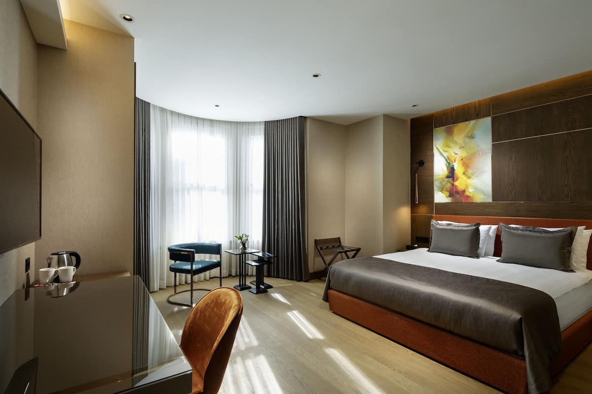 A modern hotel room with a large bed, a desk with a chair, a small table with a chair, and a large window with curtains.