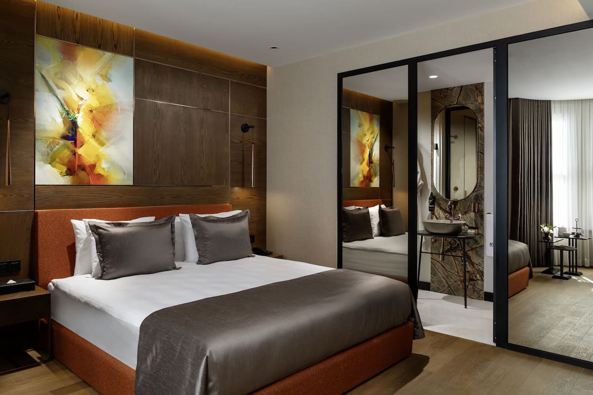 A modern hotel room with a large bed, wooden paneling, abstract art, a bathroom with a marble wall, and a sliding door leading to another room.