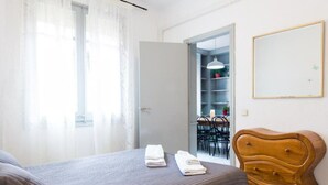 Une chambre à coucher comprenant un lit, une commode en bois, une porte donnant sur un coin repas et un tableau encadré accroché au mur.