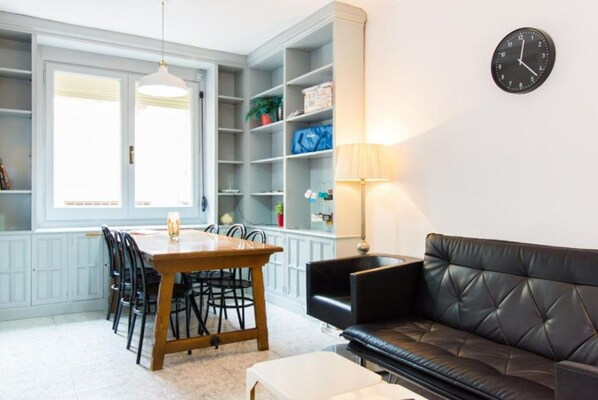 A modern living room with a dining area, a black leather sofa, a wooden dining table with chairs, and built-in shelving with various items.