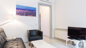Un salon avec un canapé, un fauteuil noir, une table basse blanche, un téléviseur posé sur un meuble et un tableau représentant un paysage urbain accroché au mur.