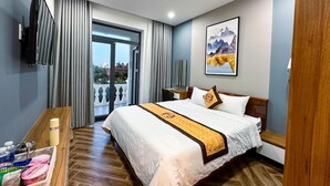 Une chambre d’hôtel avec un grand lit, une table de chevet en bois où sont posés les articles de toilette, une vue sur l’extérieur grâce à une porte coulissante, et un tableau accroché au mur.