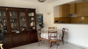 A dining area with a round table and chairs, a wooden cabinet with glass doors, and a tiled backsplash in the kitchen area. - Logement Entier : Appartement - Antibes, France (Antibes)
