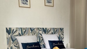 A neatly made bed with a patterned headboard, two pillows with the word "Bonheur," a rolled-up towel, and a blue towel on a white bedspread.