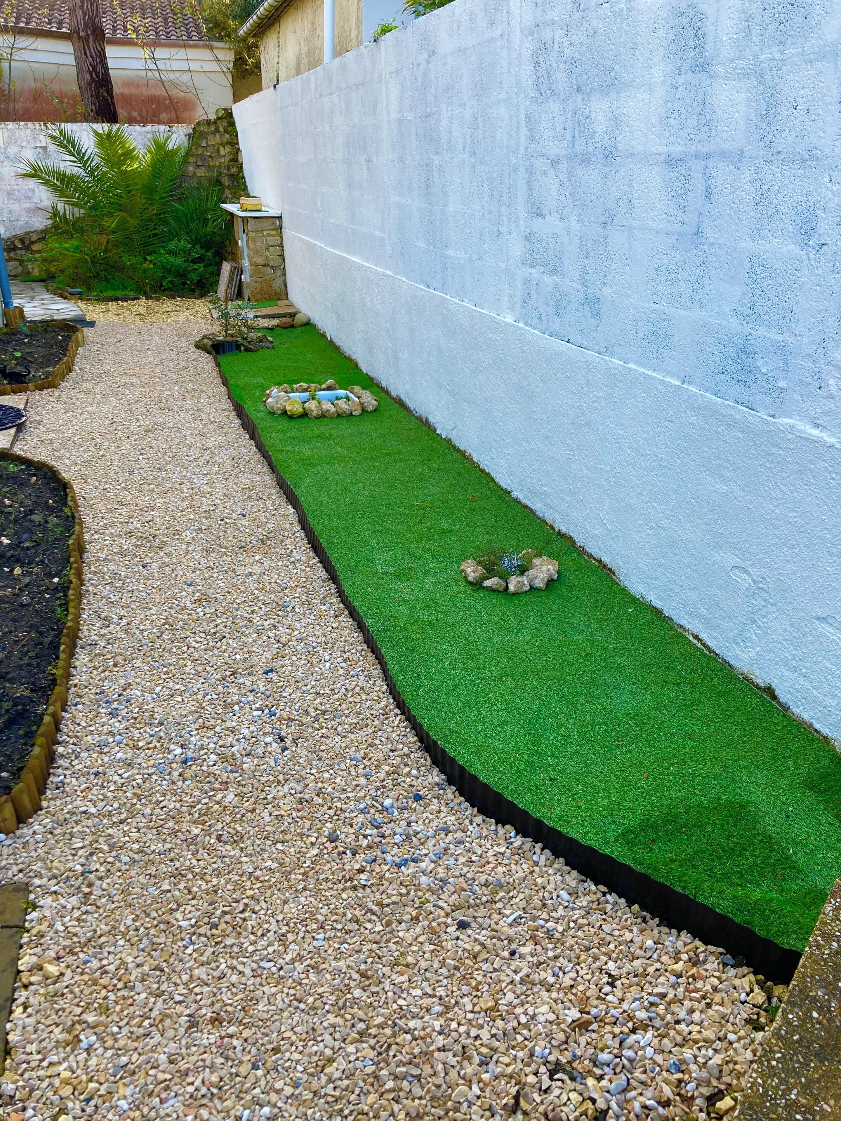 Ein Gartenbereich mit Kunstgrasanlage, Kieselsteinen und einer weißen Mauer.