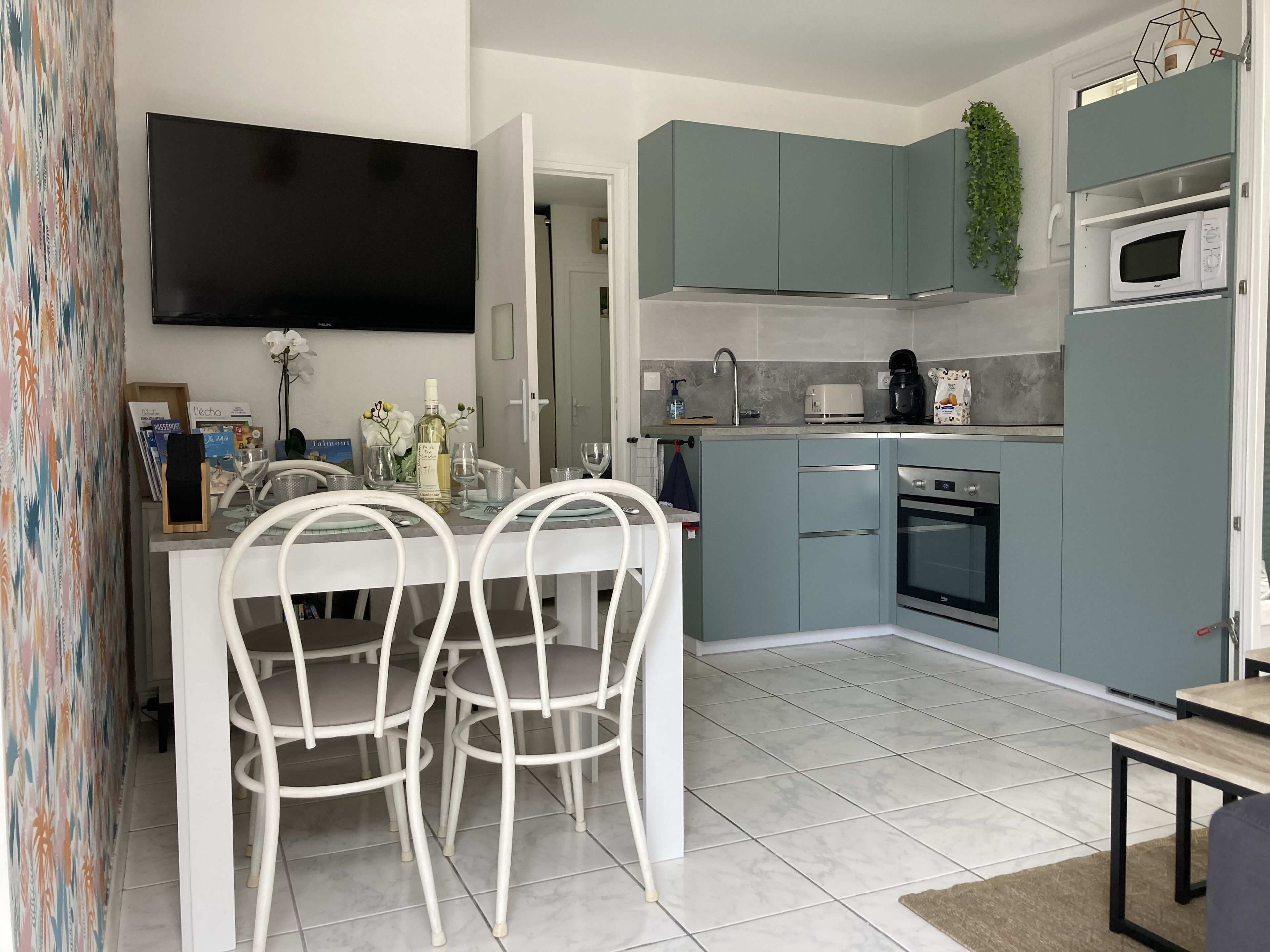 A modern kitchen with teal cabinets, a built-in oven, and a microwave. There is a dining area with white chairs and a table.
