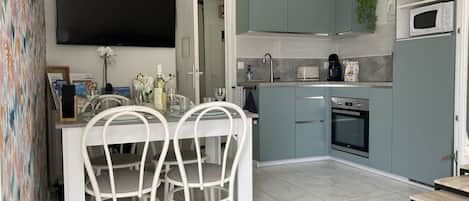 A modern kitchen with teal cabinets, a built-in oven, and a microwave. There is a dining area with white chairs and a table.