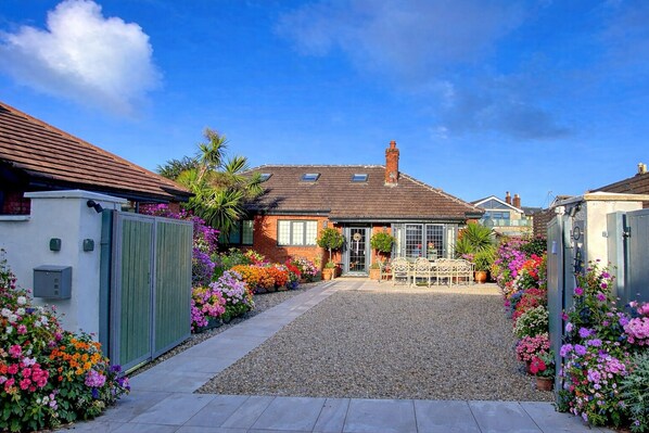 A residential property with a gravel driveway, a front garden with colorful flowers, a wooden bench, and a brick house with a tiled roof.