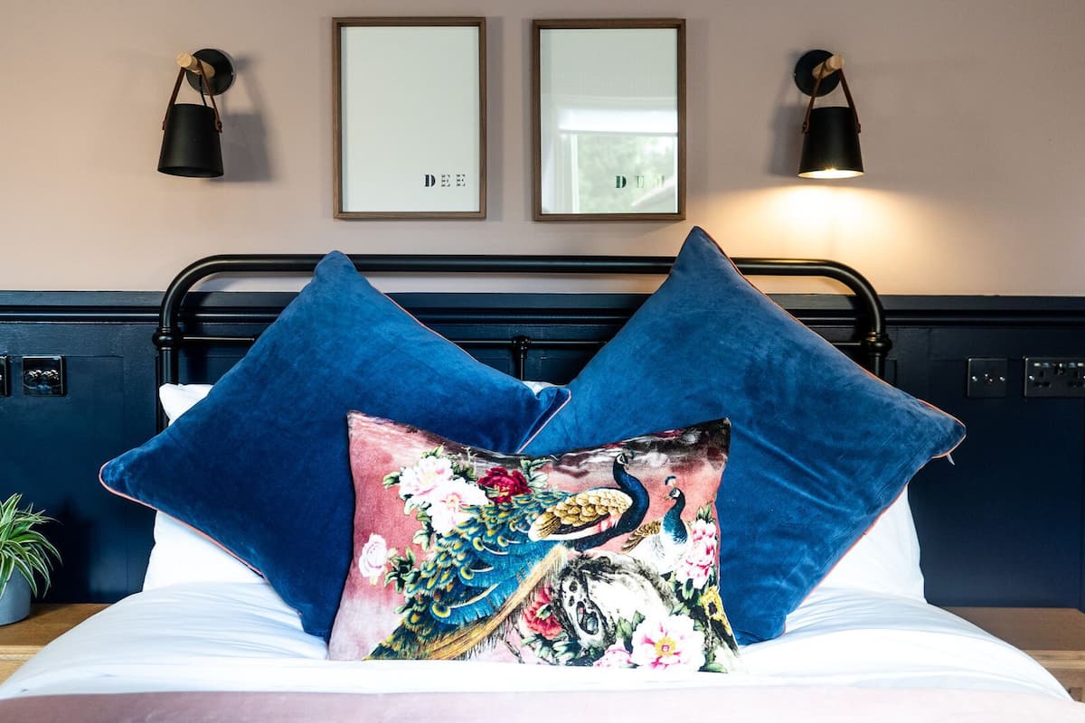 A bed with two blue pillows and a decorative pillow with a peacock design. There are two wall-mounted lamps and two framed pictures above the bed.