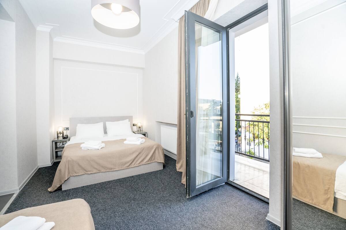 A hotel room with a bed, a small table with a lamp, a glass door leading to a balcony, and a second bed.