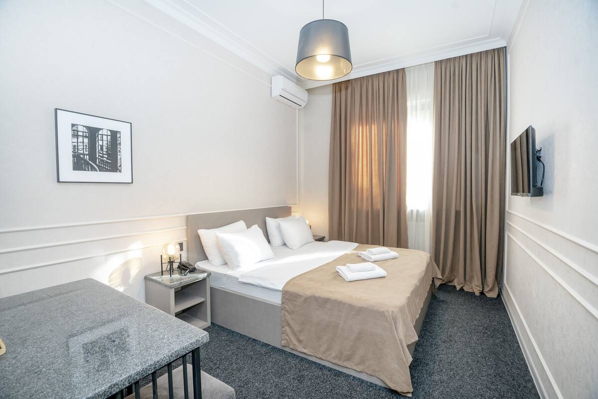 A hotel room with a bed, bedside table, lamp, air conditioning unit, and a television on the wall.