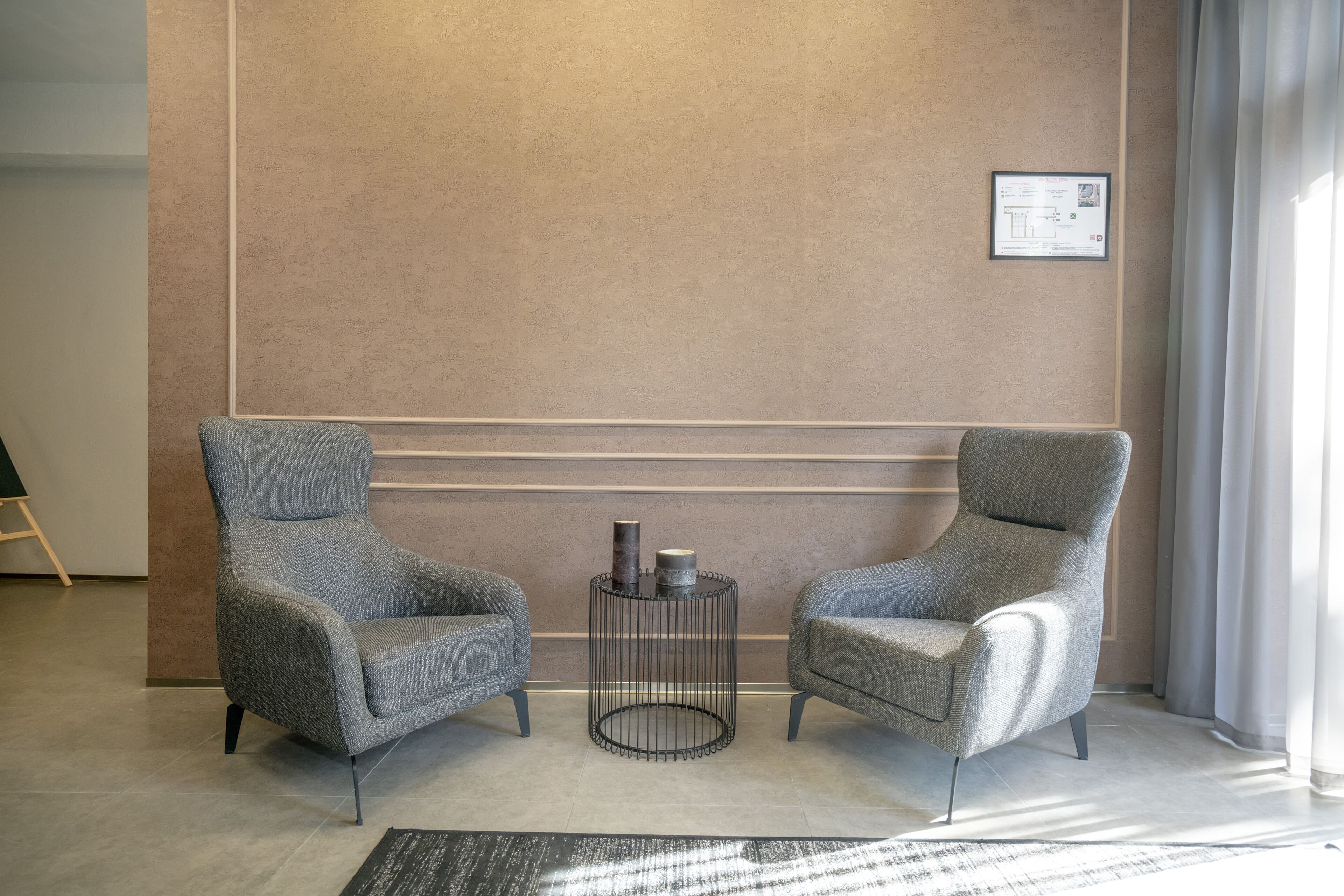 Two grey armchairs facing each other with a small round table between them, a framed certificate on the wall, and a large window with curtains.
