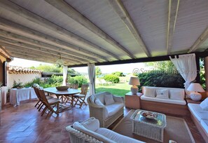 Relax in a serene outdoor space with a round table, wicker sofa, and cushioned bench amidst lush surroundings.