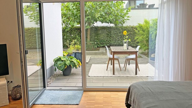 Une chambre avec une porte-fenêtre coulissante donnant sur une terrasse. La chambre est équipée d’un lit, d’une télévision et d’une petite table sur laquelle est posé un vase de fleurs.