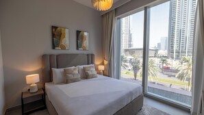 Ein modernes Hotelzimmer mit einem großen Bett, zwei Nachttischlampen und Blick auf hohe Gebäude und Palmen durch ein großes Fenster.