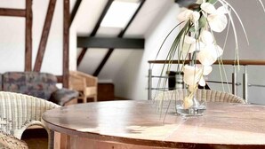 A round wooden table with a clear glass vase containing white flowers and green leaves. The room features exposed wooden beams and a sloped ceiling, suggesting a loft-style interior. - *Schwarzwald-Haus mit Sauna* (Elzach)