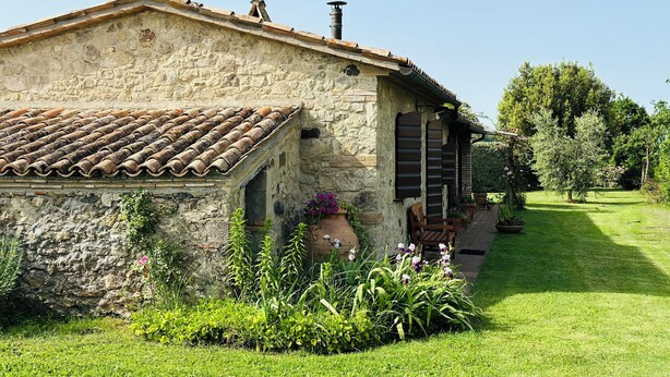 A rustic stone building with a tiled roof, a small porch with a chair, and a garden with various plants and flowers. - Milva's Nest Countryside (Umbria)