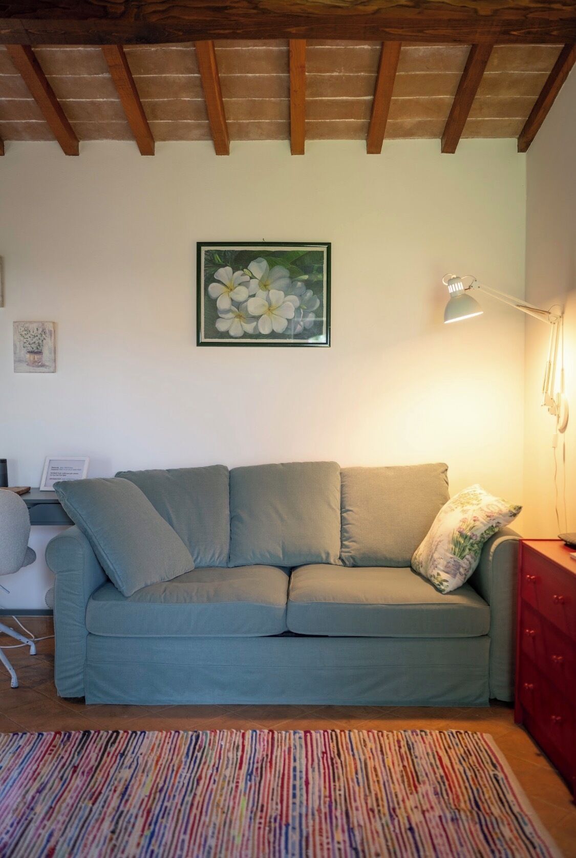 Un salon avec un canapé, un tableau floral encadré, un bureau avec une lampe et un tapis coloré.