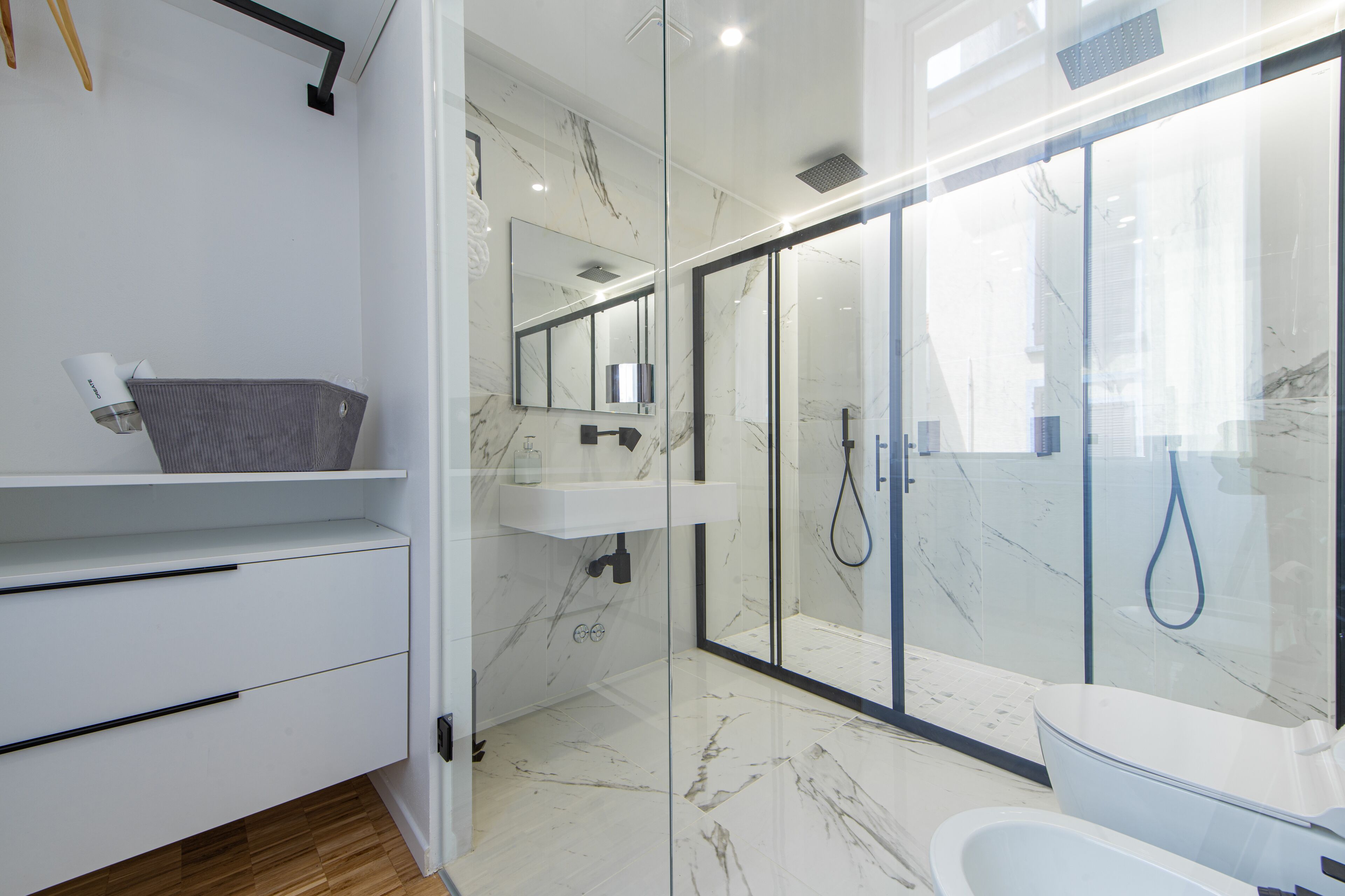 Un baño moderno con ducha de vidrio, lavamanos blanco y tocador con cajones.
