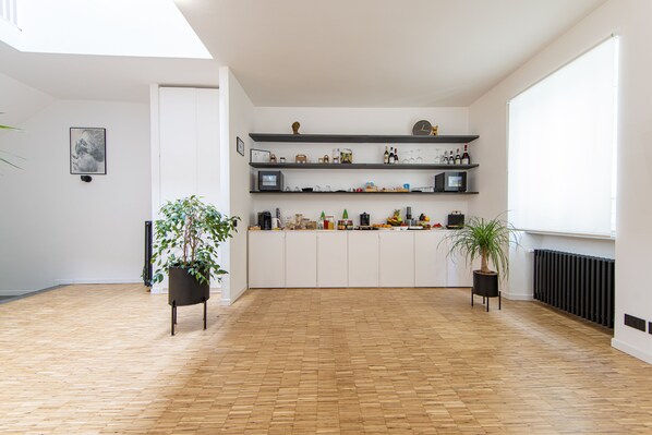 A spacious room with a wooden floor, white walls, and a black-framed TV on a shelf. There are two potted plants and a radiator on the wall.