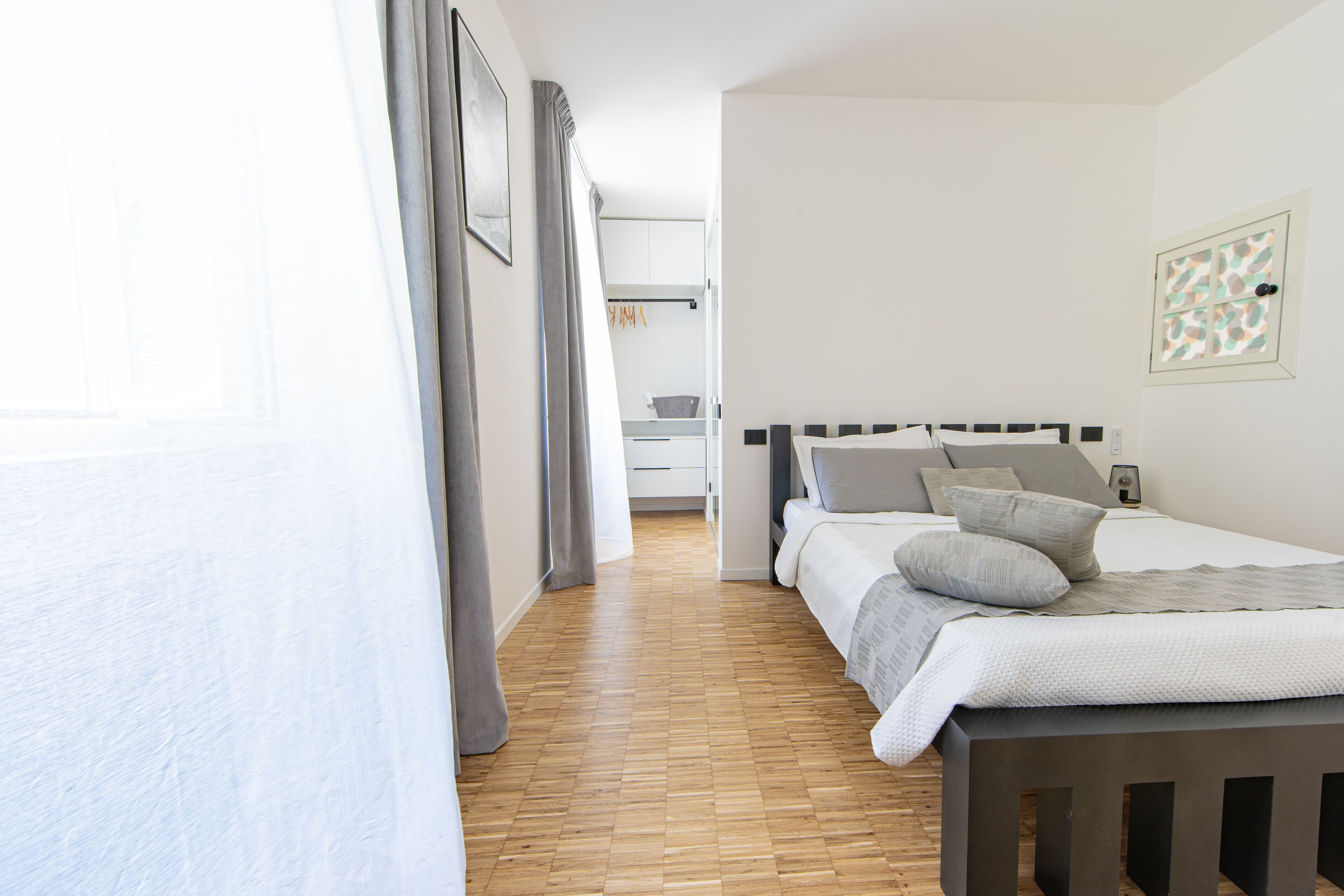 A bedroom with a wooden floor, a bed with grey bedding, a bedside table, and a window with curtains.