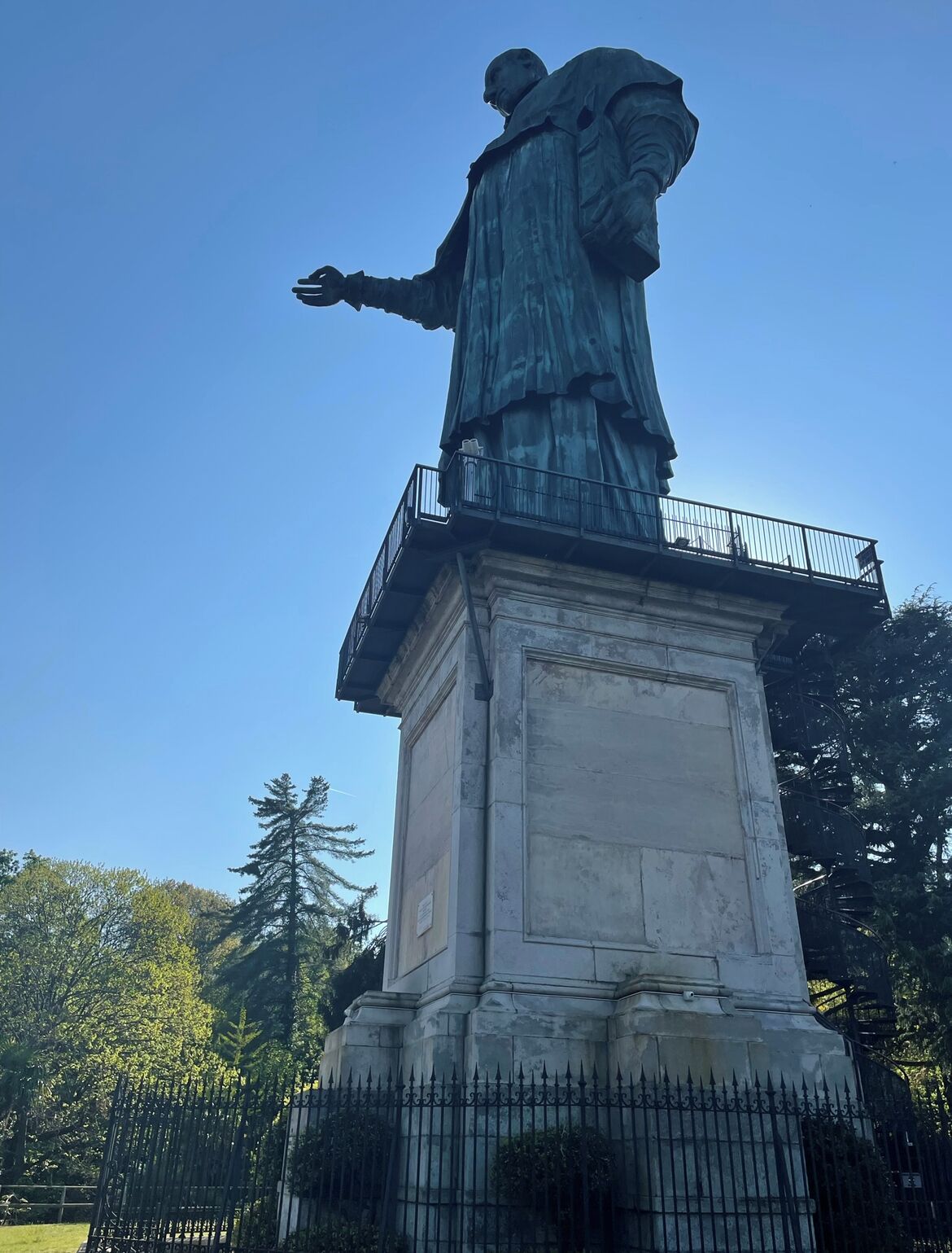 A statue on a pedestal with a balcony, surrounded by a fence and trees.