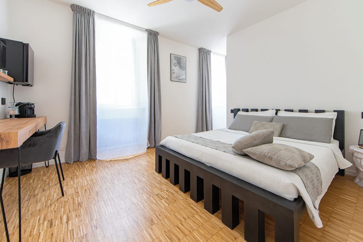 Une chambre moderne avec un plancher en bois, un lit avec une literie grise, un bureau avec une chaise, une télévision et une fenêtre avec des rideaux.