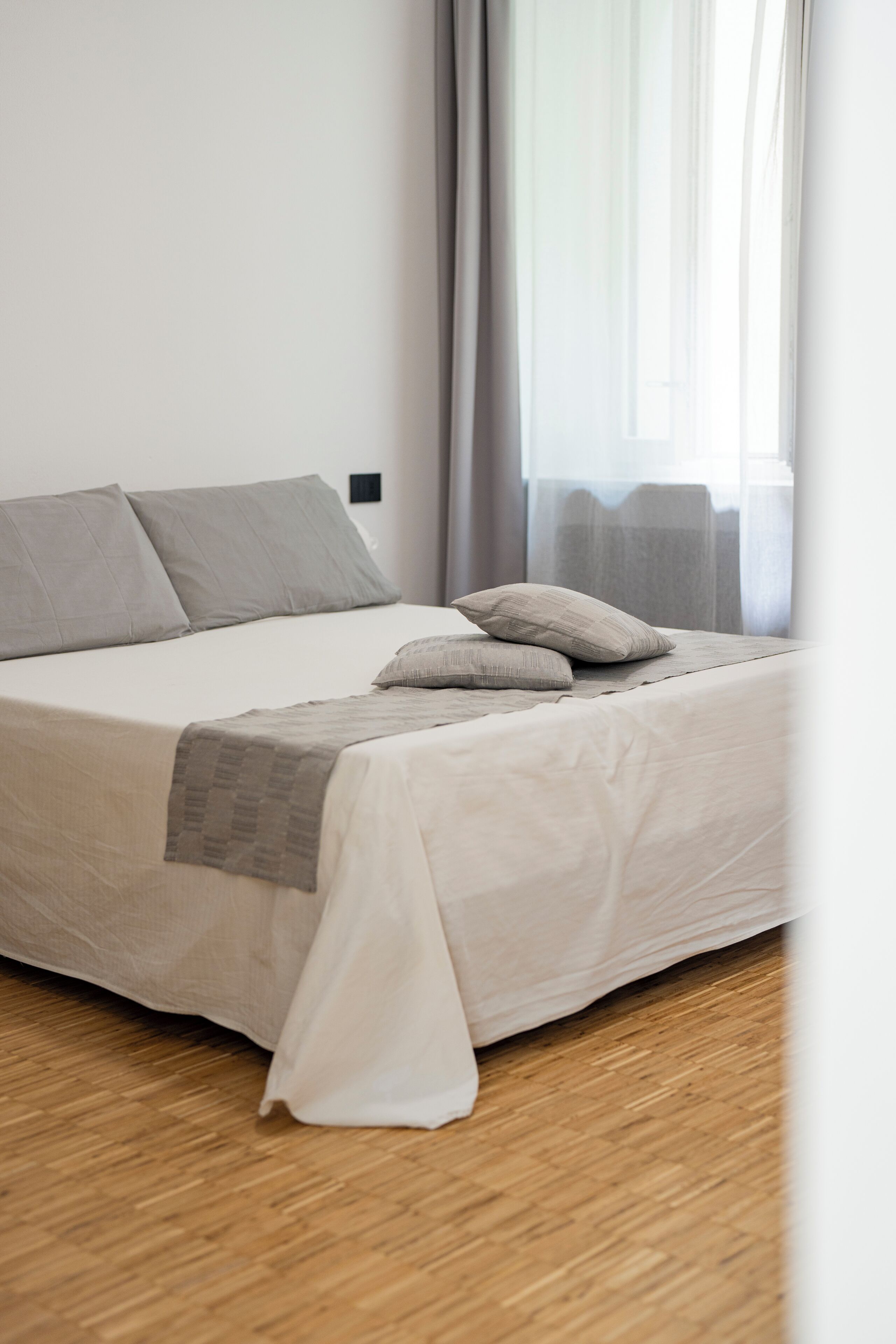A neatly made bed with a white and gray duvet, a gray throw blanket, and two pillows. The room has a wooden floor and a window with sheer curtains.