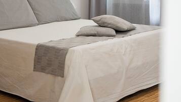 A neatly made bed with a white and gray duvet, a gray throw blanket, and two pillows. The room has a wooden floor and a window with sheer curtains.