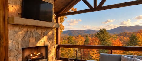 A stone fireplace with a TV above it, a wooden deck with outdoor furniture, and a view of autumn foliage and mountains.