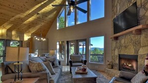 A spacious living room with a high ceiling, large windows, a stone fireplace, and a comfortable seating area.