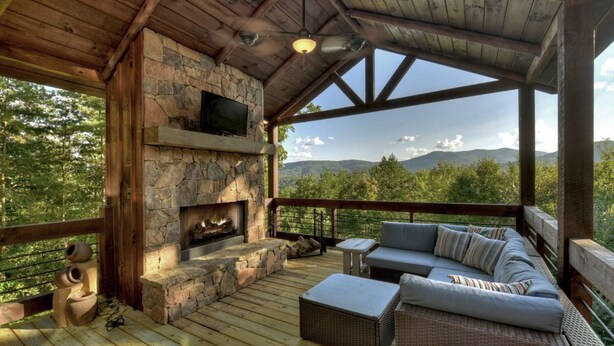 A covered outdoor area with a stone fireplace, a television mounted above it, and a seating area with cushions.