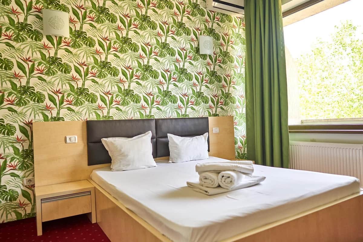 A hotel room with a bed, two pillows, a nightstand, a lamp, and a window with curtains.