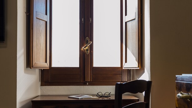 A wooden desk with a chair, a window with wooden shutters, and a pair of glasses on the desk.