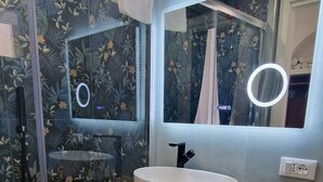 A modern bathroom with a glass shower enclosure, a white sink on a wooden vanity, and a mirror with a circular light. - B (Napoli)