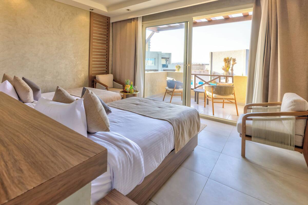 A hotel room with two beds, a balcony with a chair and table, and a view of the city.