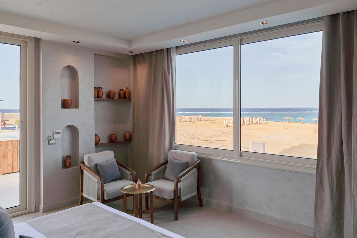 A room with a large window overlooking a beach, two armchairs, a small table, and shelves with decorative items.