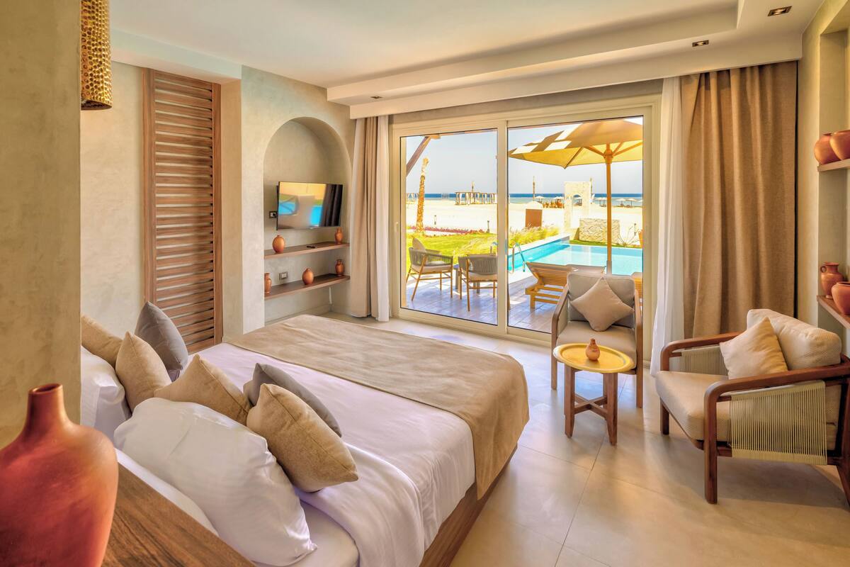 A hotel room with a large bed, a TV, a small table, and a view of the beach and pool.