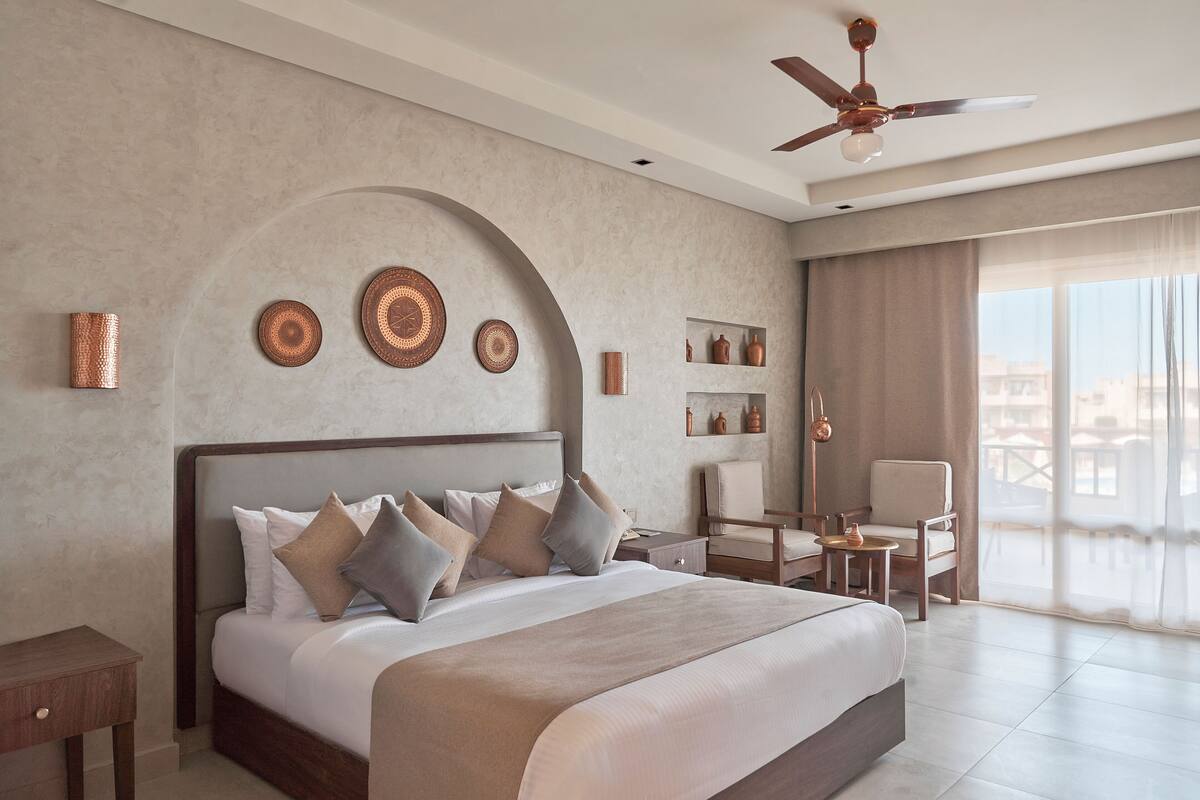 A modern hotel room with a large bed, a desk, a chair, and a balcony with a view.