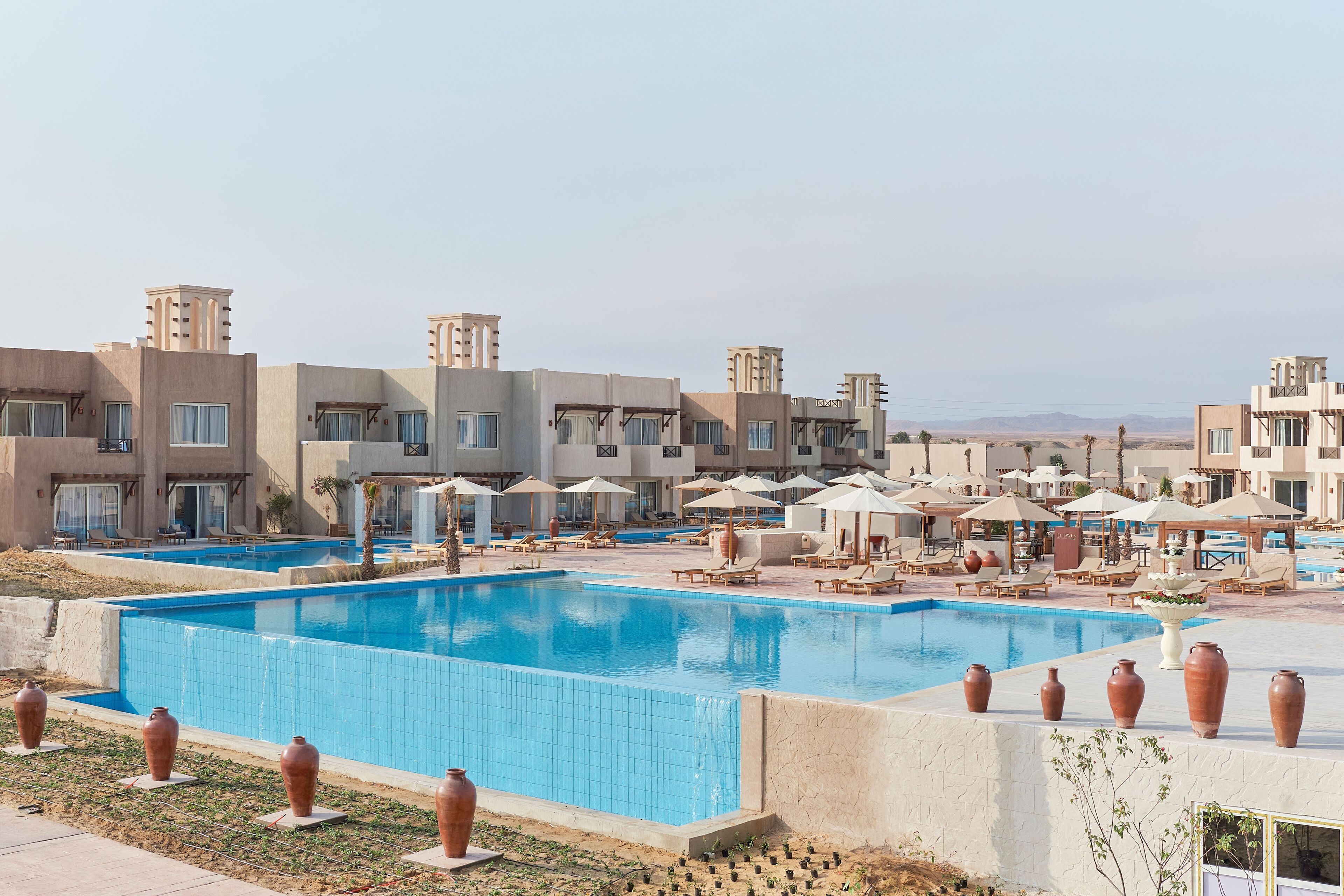 Área de piscina do resort com várias espreguiçadeiras e guarda-sóis, rodeada por edifícios com arquitetura uniforme.